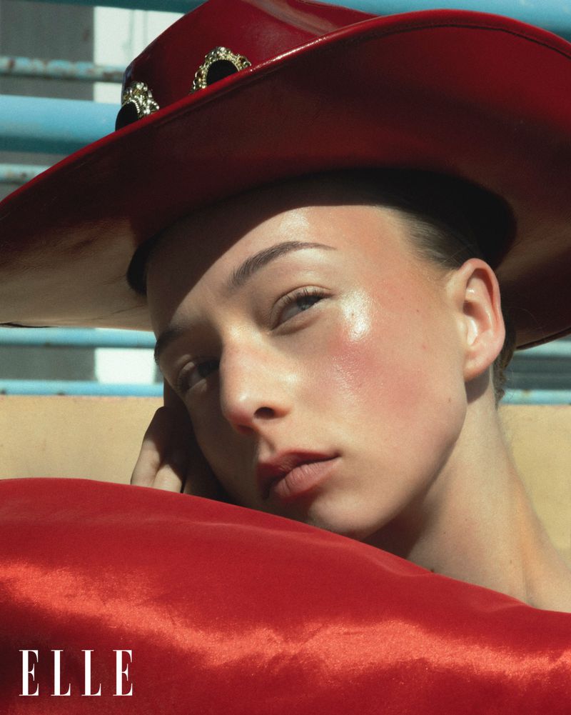 Close-up portrait of a young adult woman wearing a shiny red cowboy hat and matching satin shawl, bathed in bright sunlight.