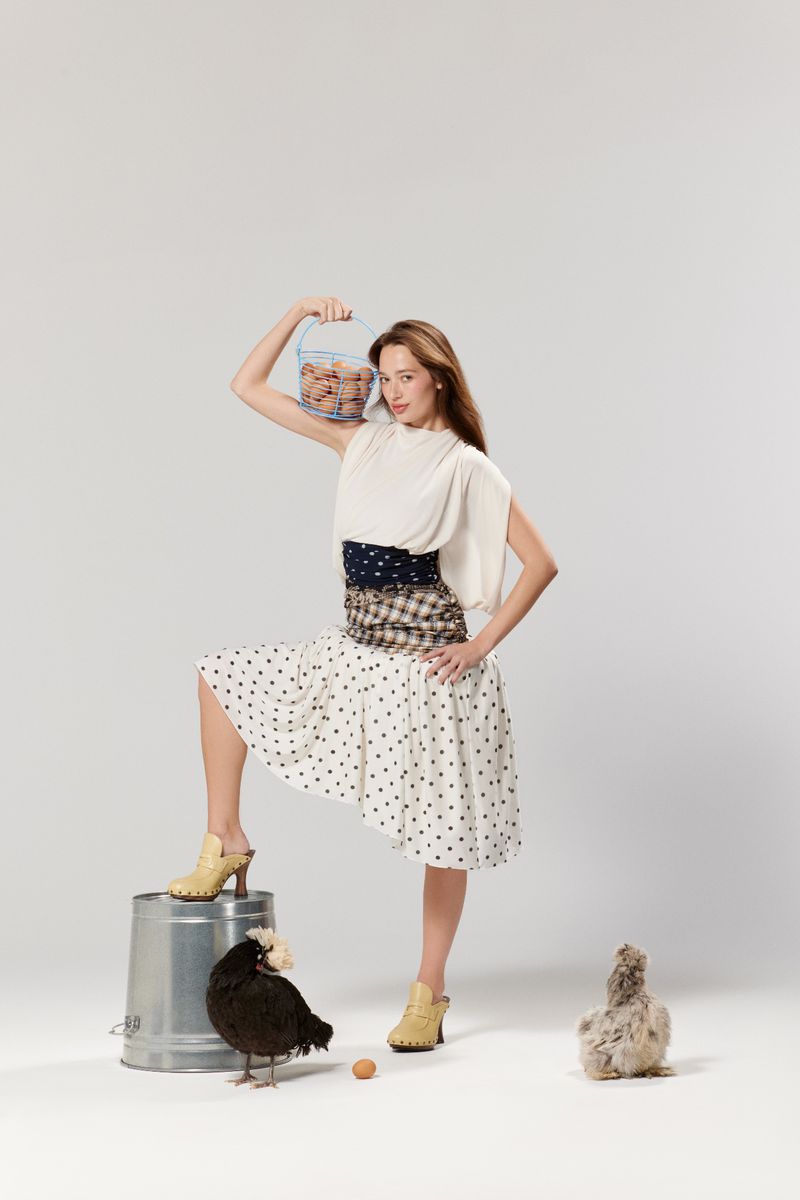 Elegant woman in patterned dress poses with a wire basket of eggs, standing near a black hen and chick in a bright photo studio.