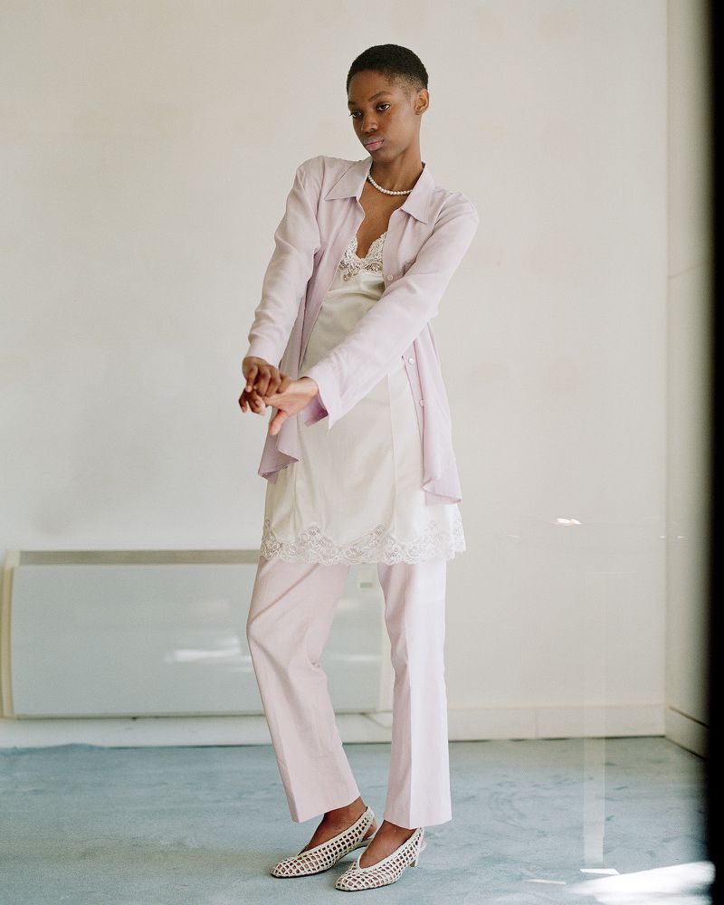 A young Black woman in a pastel lavender linen shirt and pants, over a white lace slip dress, poses in front of an off-white wall.