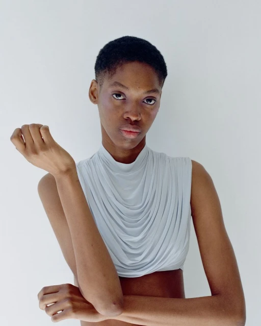 Close-up studio shot of a young adult woman with a short haircut wearing a draped, light-blue top against a white background.