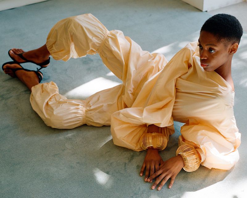 Model lying on a pale green carpet wearing a light yellow parachute jumpsuit and large gold cuffs