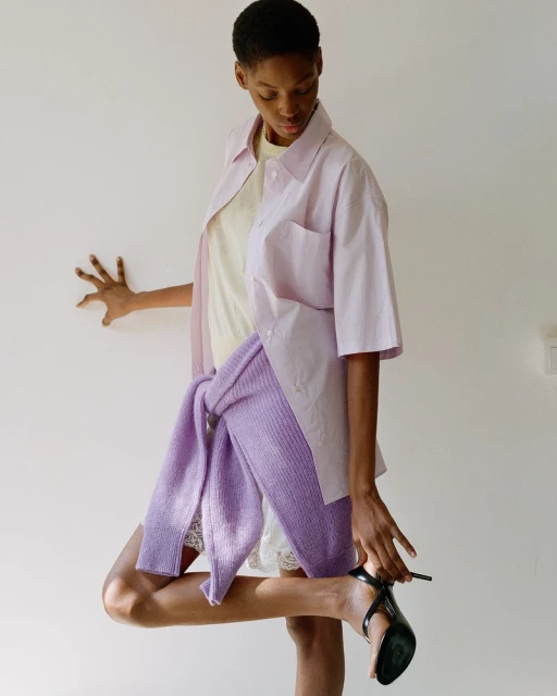 Woman in pink shirt and sweater putting on black high-heeled shoe against white wall. Fashion editorial shot indoors.