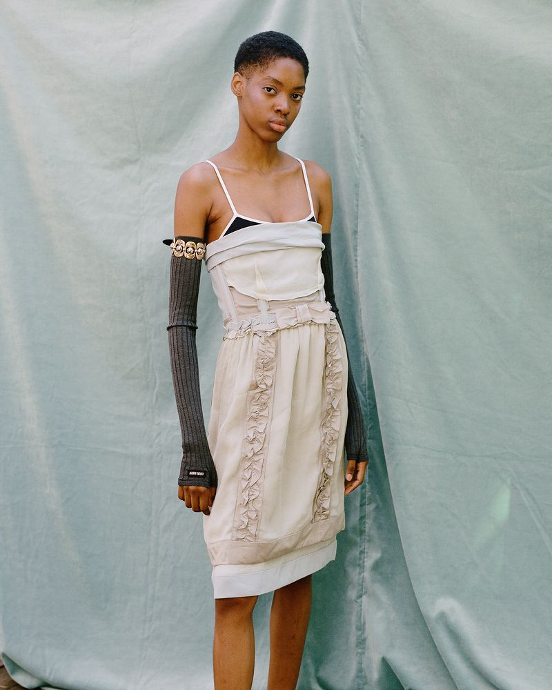An androgynous fashion model poses outdoors in a light colored dress with long, dark grey arm sleeves against a teal backdrop.