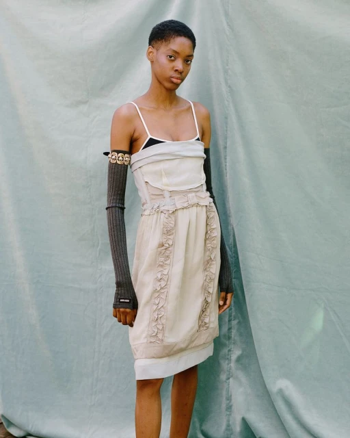 An androgynous fashion model poses outdoors in a light colored dress with long, dark grey arm sleeves against a teal backdrop.