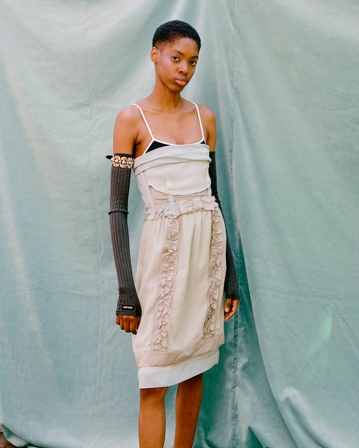 Model with short dark hair wearing a layered beige dress and arm warmers against a textured green backdrop for M Le magazine du Monde