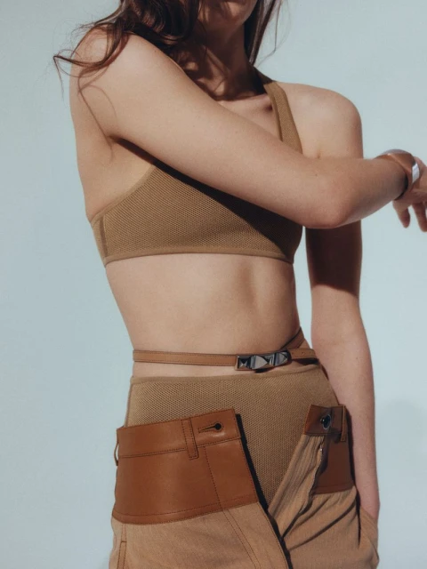 A slender woman with tan skin poses indoors wearing a tan-colored bra top and tan pants with leather-trimmed accents, captured in a studio shot.