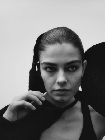 Striking monochrome portrait of a young adult woman with slicked-back hair wearing a black top and textural head covering against a white backdrop.
