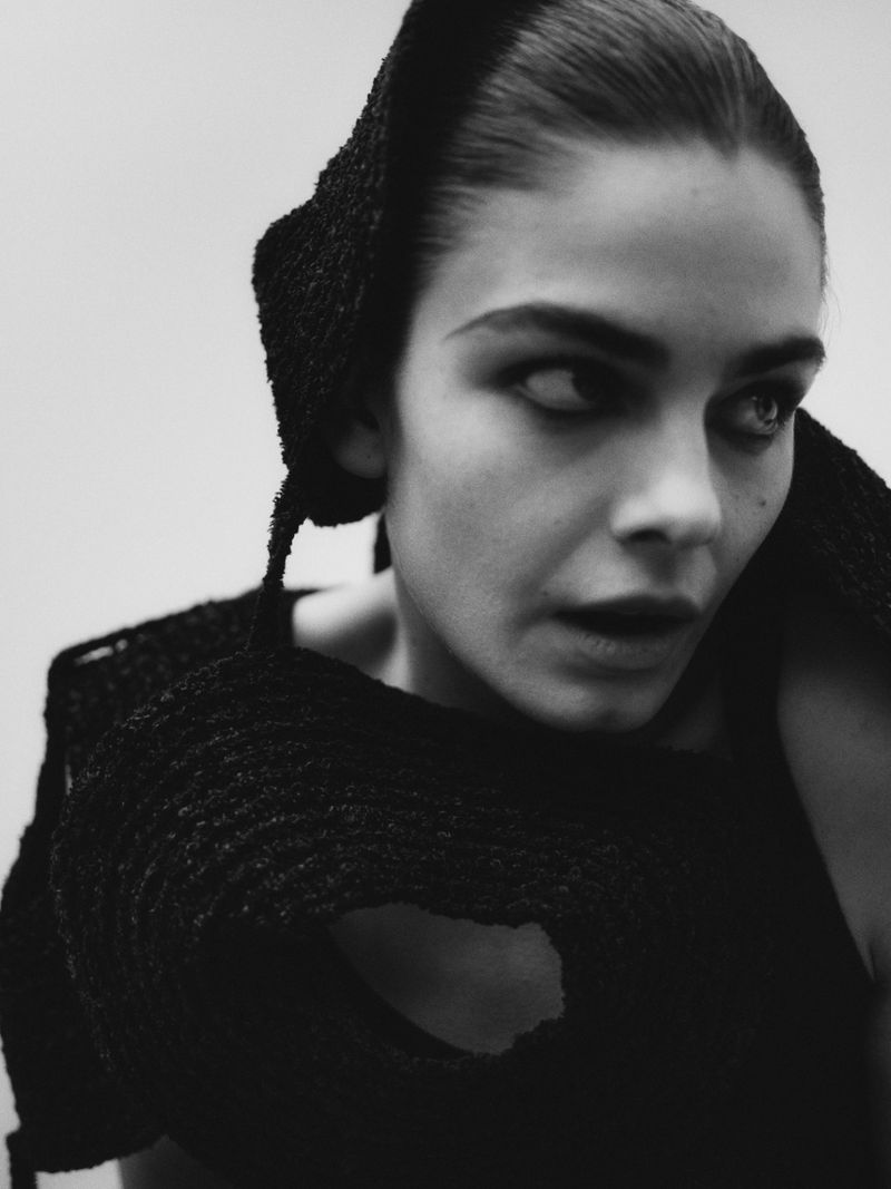 Monochrome portrait of a fair-skinned woman with dark hair wearing textured black clothing and head covering, looking to the side.