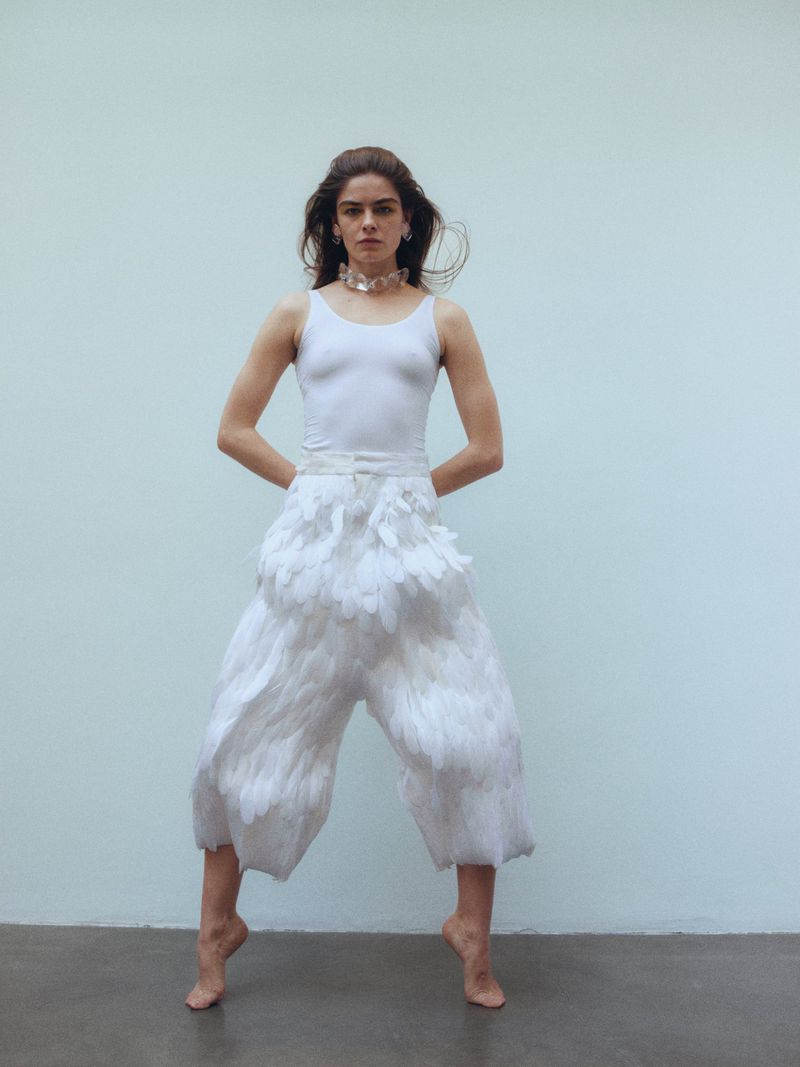 Young woman strikes a pose in a white top and feather pants against a plain backdrop, capturing a unique blend of fashion and art.