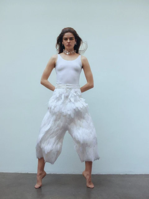 Young woman strikes a pose in a white top and feather pants against a plain backdrop, capturing a unique blend of fashion and art.