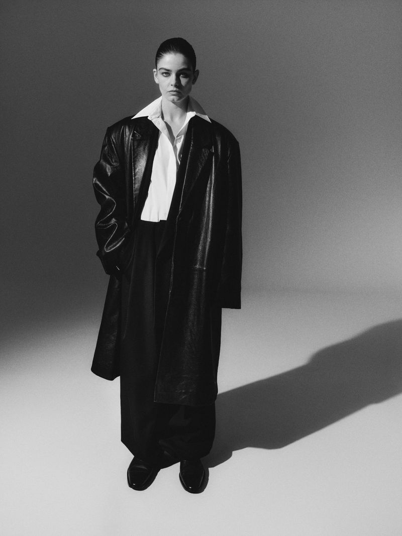 Black and white fashion shot of a woman in an oversized leather coat and dress pants, posed in a photo studio with directional lighting.