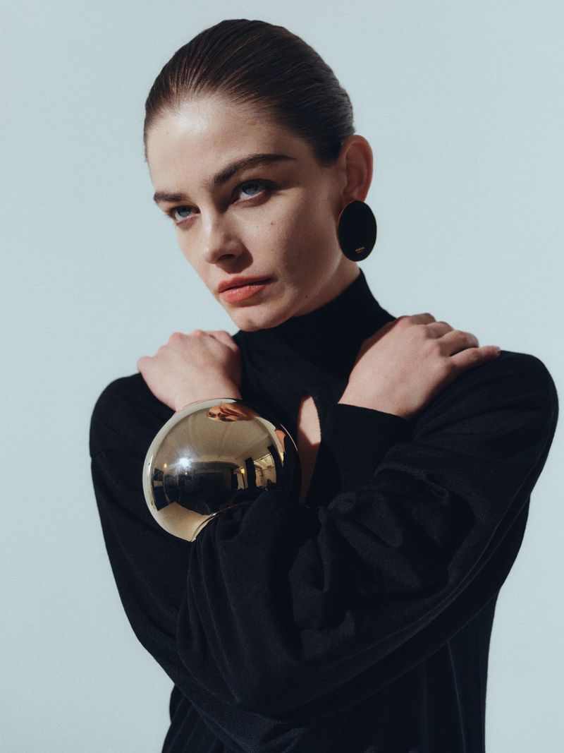A young woman with her arms crossed and wearing a black turtleneck, gold bangle, and large black earrings poses in a studio setting.