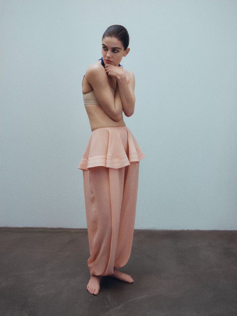 Fashion shot of a young adult woman in a peach-colored outfit posing in front of a neutral background in an indoor setting.