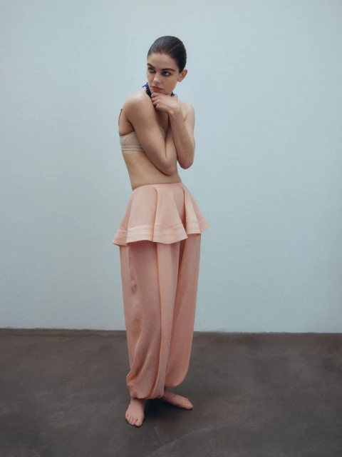 Fashion shot of a young adult woman in a peach-colored outfit posing in front of a neutral background in an indoor setting.