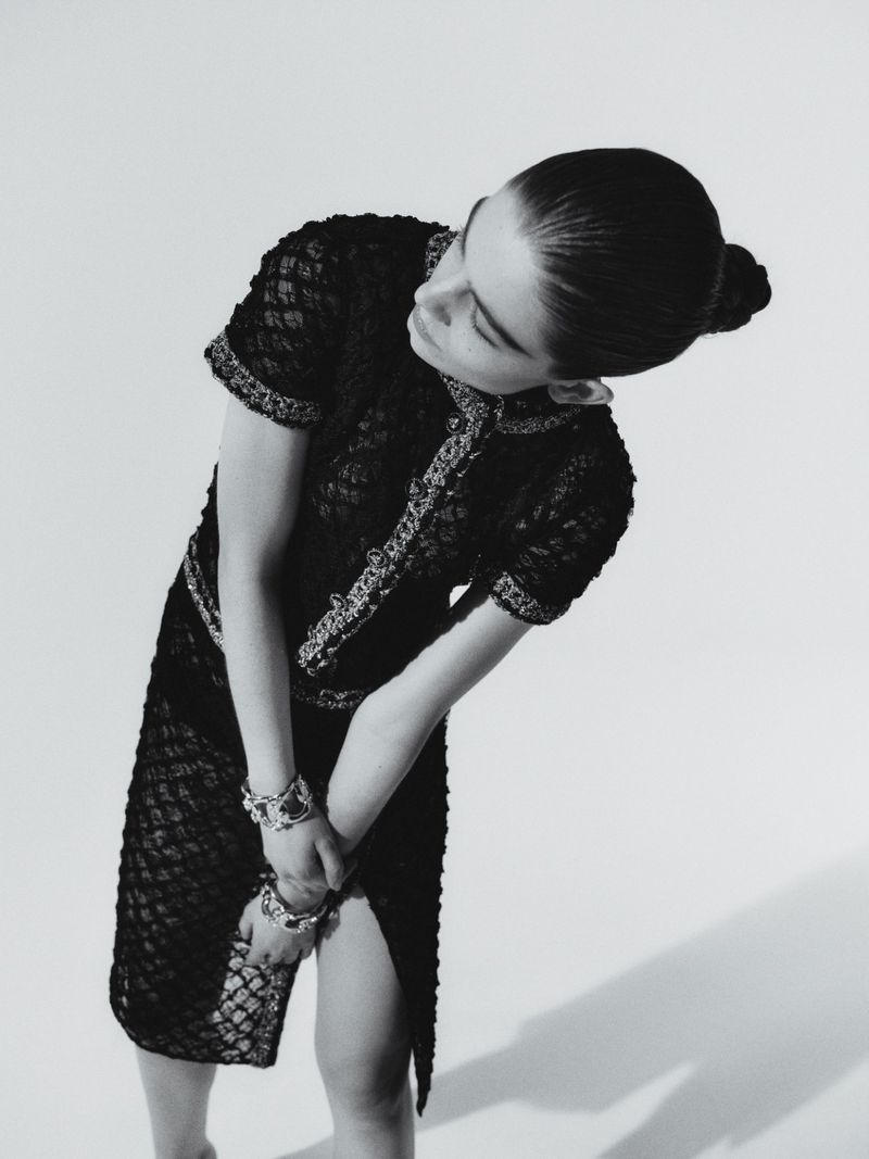 Monochrome fashion shot of a female adult model in an ornate black lace dress, posing indoors with hands clasped and head tilted.
