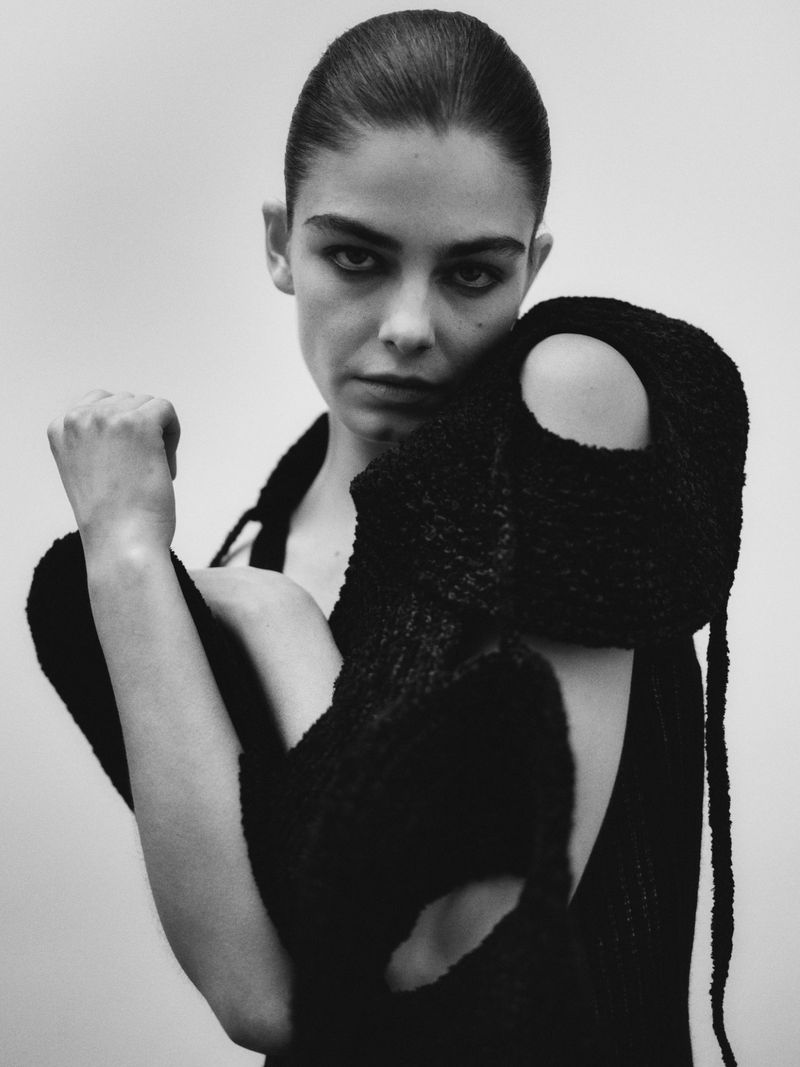 Monochrome portrait of a woman with dark hair and a dark open-back knit garment with cutout details, posing against a light backdrop.