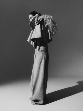 Fashion shot of a woman wearing wide-leg jeans and a denim top with fringe detail, standing in a photo studio against a grey backdrop.