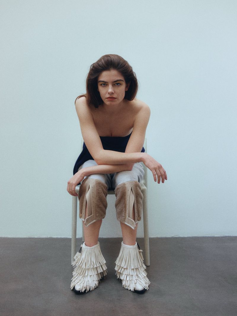 Woman in fashion footwear sits on chair against white wall. Stylish look with unique pants and fringed boots in a minimalist setting.