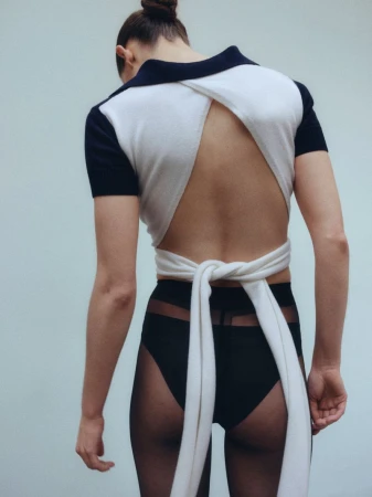 Back view of a female fashion model wearing a white open-back wrap top, black undergarments, and sheer tights, posing against a pale backdrop.