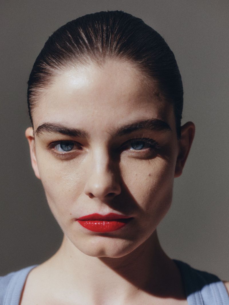 Close-up portrait of a beautiful young woman with red lipstick and blue eyes, set against a neutral background with natural lighting.