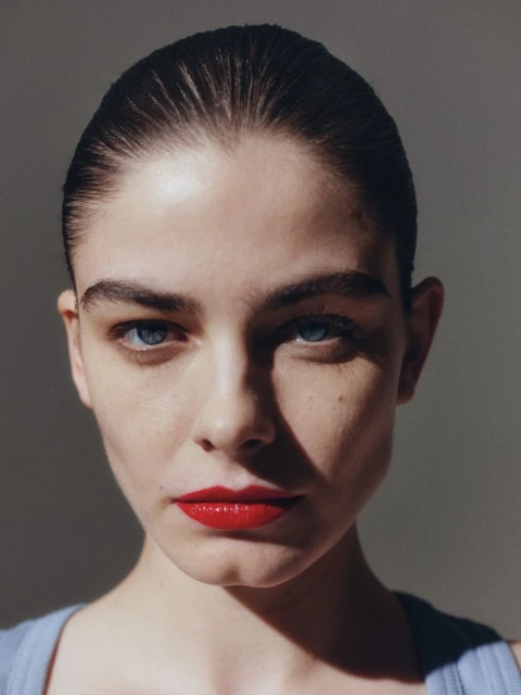 Close-up portrait of a beautiful young woman with red lipstick and blue eyes, set against a neutral background with natural lighting.