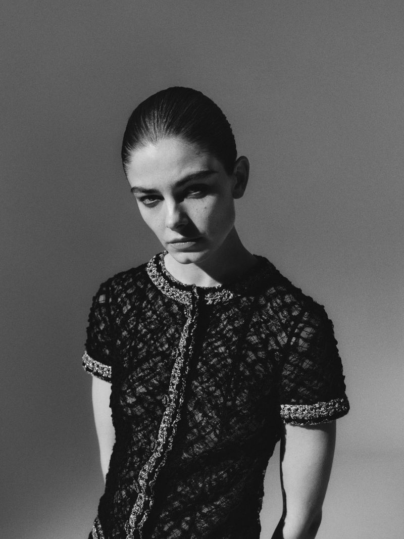 Monochrome portrait of a woman in a black, textured top with embellished trim, set against a muted gray background with soft lighting.