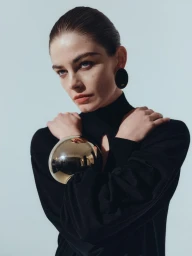 Model wearing a black top with large gold sphere bracelet and statement earrings against a seamless white background.