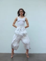 Model standing on tiptoe wearing a white tank top and feather-embellished trousers against a neutral background