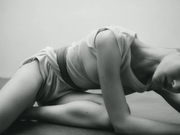 A person in a light-colored outfit bends forward with one leg extended in a minimalist, black-and-white indoor photo.