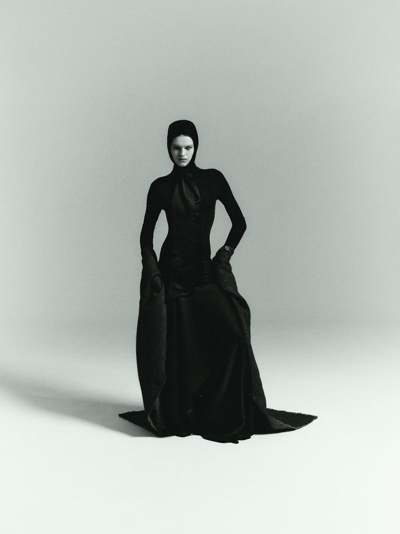 Model in black hooded dress and long dark coat posing against a pale grey studio background by Janne Rugland