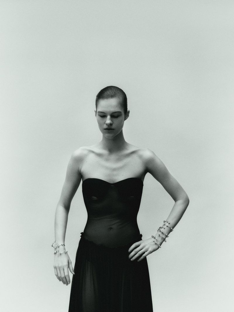 Model with a buzzcut wearing a black strapless dress against a pale grey seamless studio background.