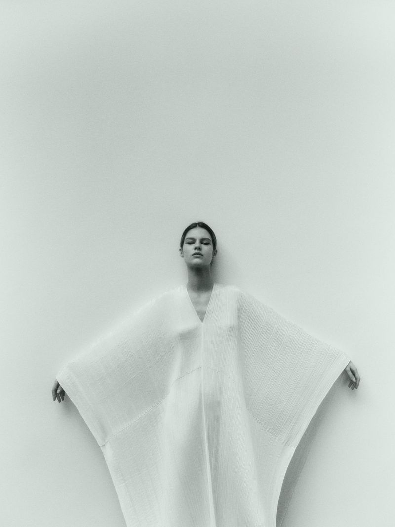A woman in a white dress with outstretched arms stands against a plain white wall in a studio-lit fashion shot.