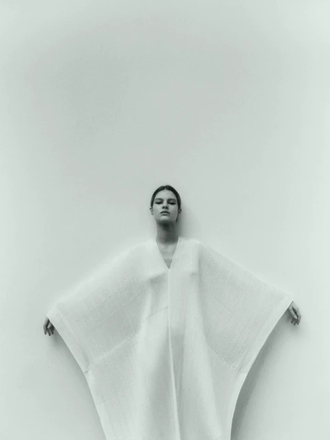 A woman in a white dress with outstretched arms stands against a plain white wall in a studio-lit fashion shot.