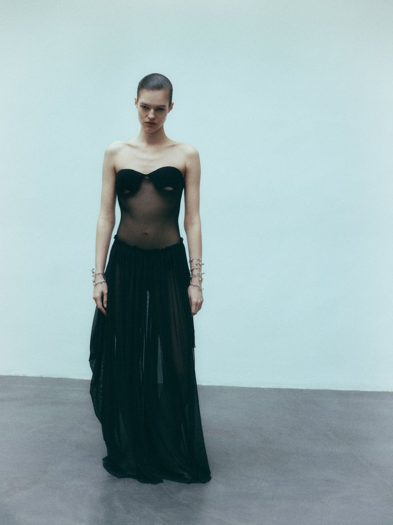 Elegant, slender female fashion model poses in a sheer black dress and silver bracelets in a bright studio environment. 