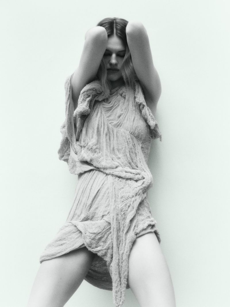 Monochrome portrait of a woman with long hair in a draped textile dress, posing against a light solid background.