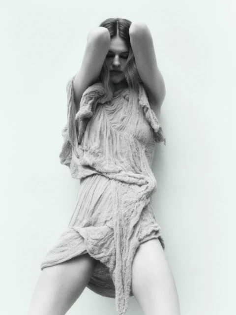 Monochrome portrait of a woman with long hair in a draped textile dress, posing against a light solid background.