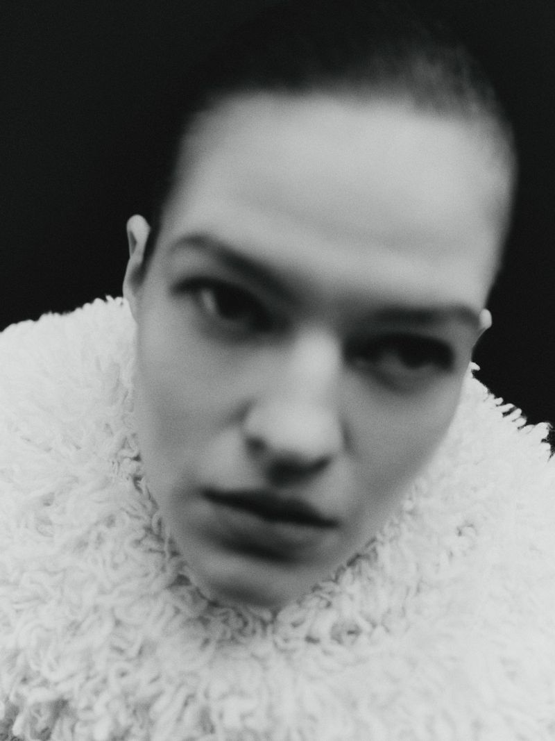 Monochrome portrait of a woman with short hair wearing a textured white coat, set against a stark black background.