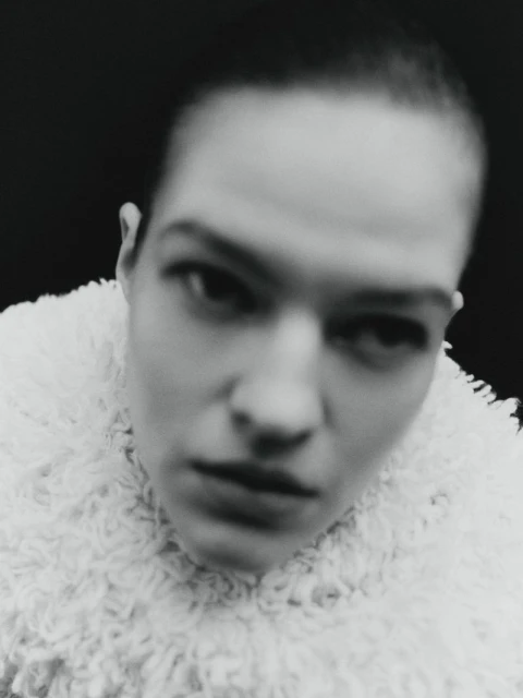 Monochrome portrait of a woman with short hair wearing a textured white coat, set against a stark black background.