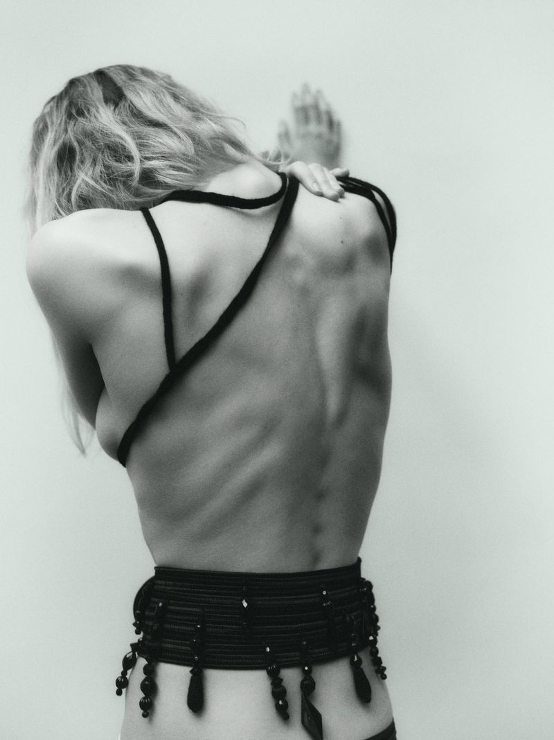 Monochrome portrait of a slender, fair-skinned adult woman, seen from the back, hands on wall, wearing black cord top and beaded belt.
