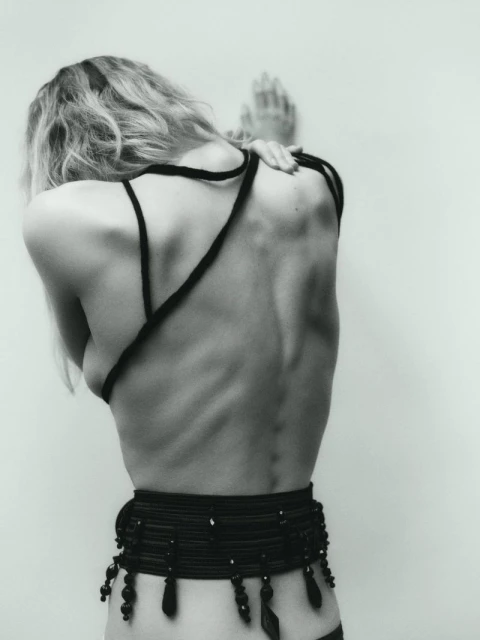 Monochrome portrait of a slender, fair-skinned adult woman, seen from the back, hands on wall, wearing black cord top and beaded belt.