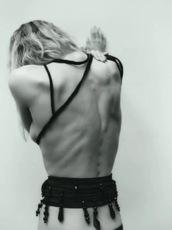 Monochrome portrait of a slender, fair-skinned adult woman, seen from the back, hands on wall, wearing black cord top and beaded belt.