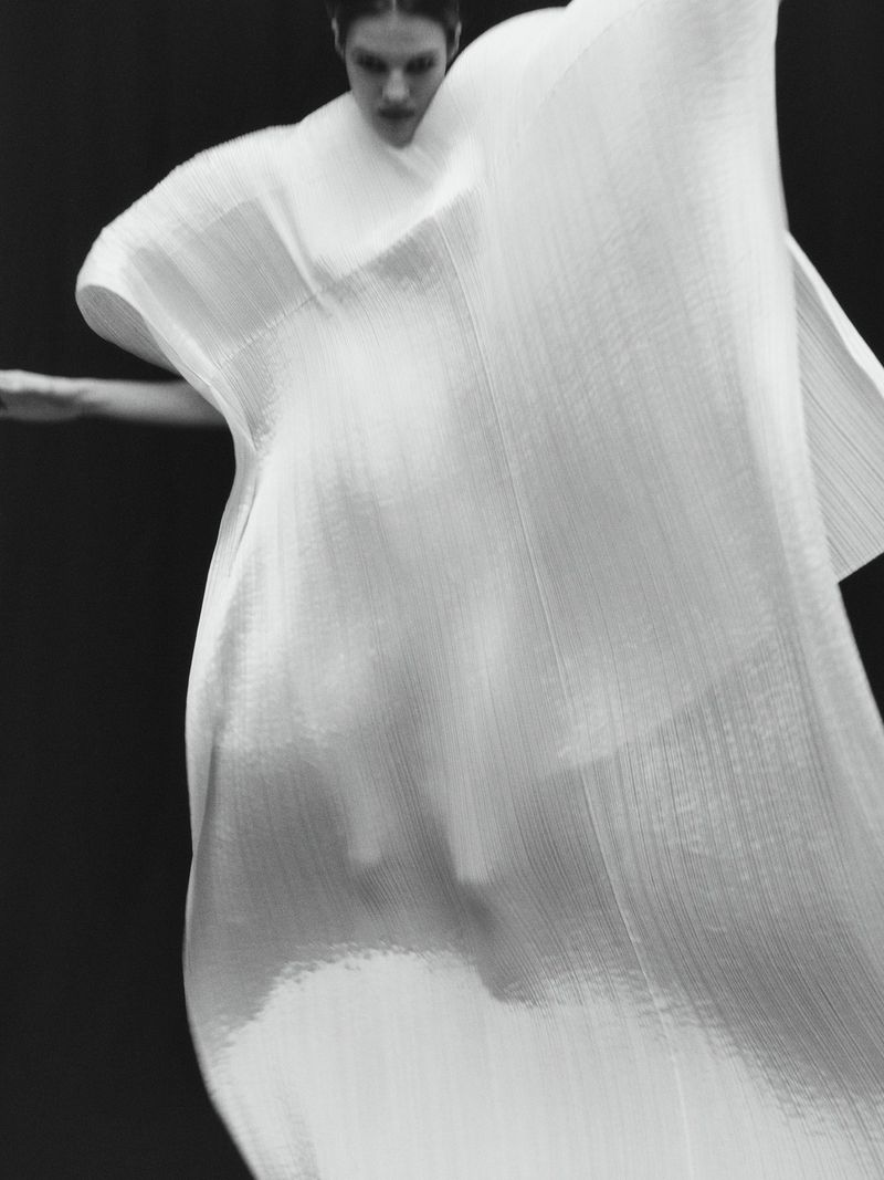 Monochrome image of a person wearing a draped, textured white dress against a black backdrop in a fashion or fine art context.