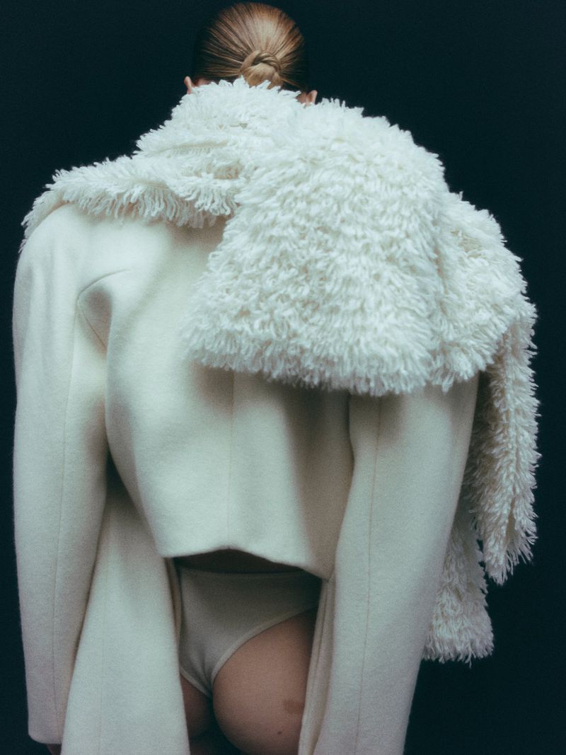 Model seen from behind wearing a white coat and fluffy white scarf against a dark background.