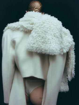 Model seen from behind wearing a white coat and fluffy white scarf against a dark background.
