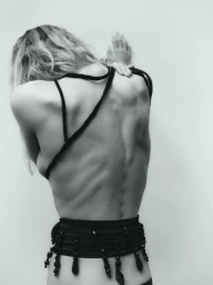 Model Angelina Artemieva posed facing a pale wall with back turned, wearing a black strap garment and beaded belt, photographed by Janne Rugland