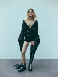Model wearing an off-the-shoulder black top, dark shorts, and tall black boots standing against a light background