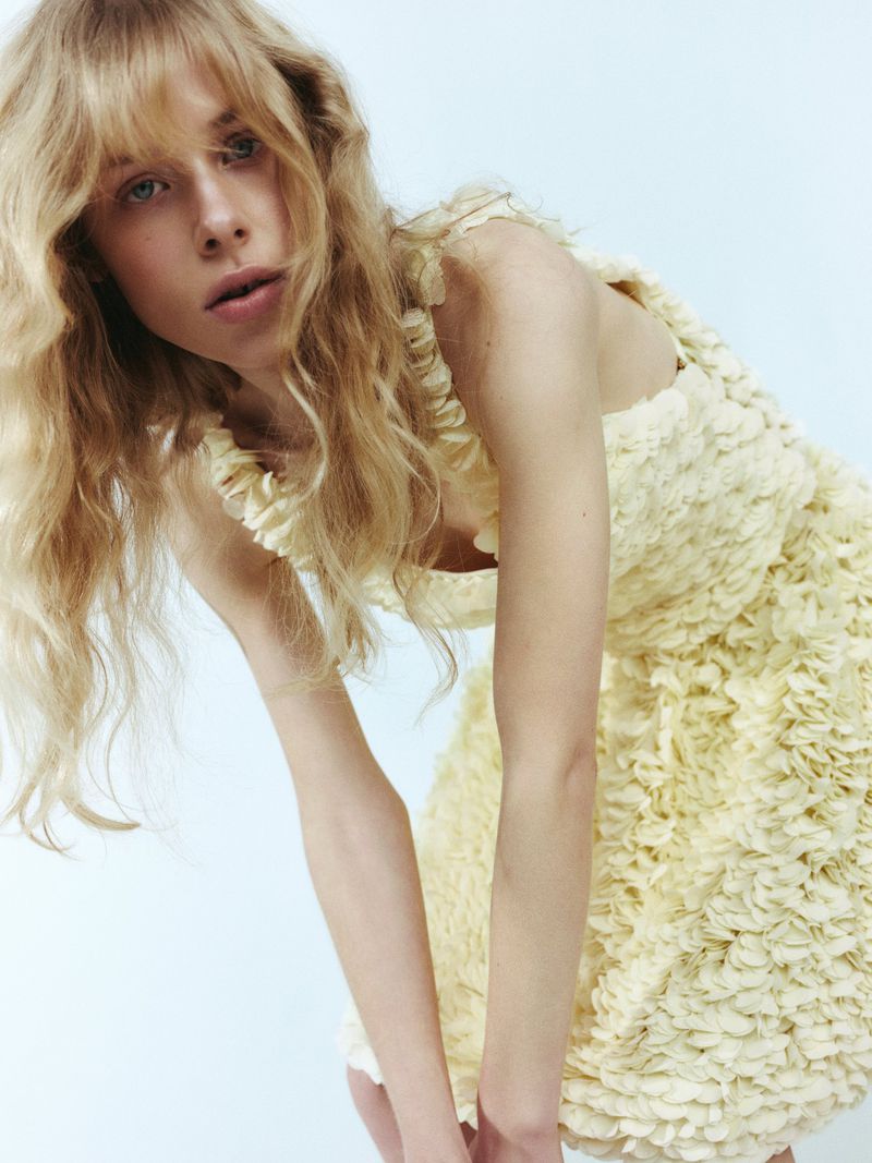 Blonde woman in a yellow textured dress looking at the camera in a studio setting. The dress has floral-like textures and ruffle details.