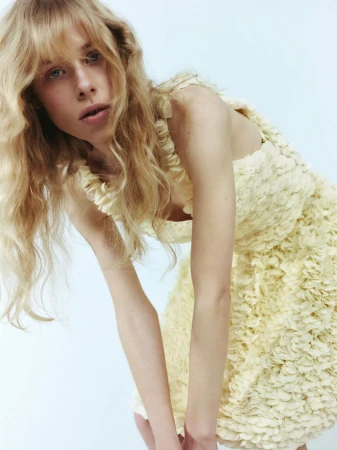 Blonde woman in a yellow textured dress looking at the camera in a studio setting. The dress has floral-like textures and ruffle details.