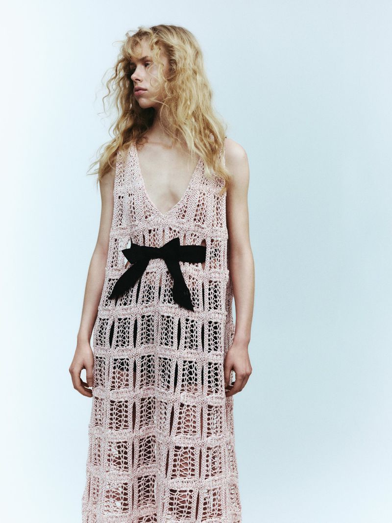 A slender adult woman with long blonde hair is posing in a pink crocheted dress with a black bow, set against a pale blue background.