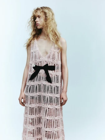 A slender adult woman with long blonde hair is posing in a pink crocheted dress with a black bow, set against a pale blue background.